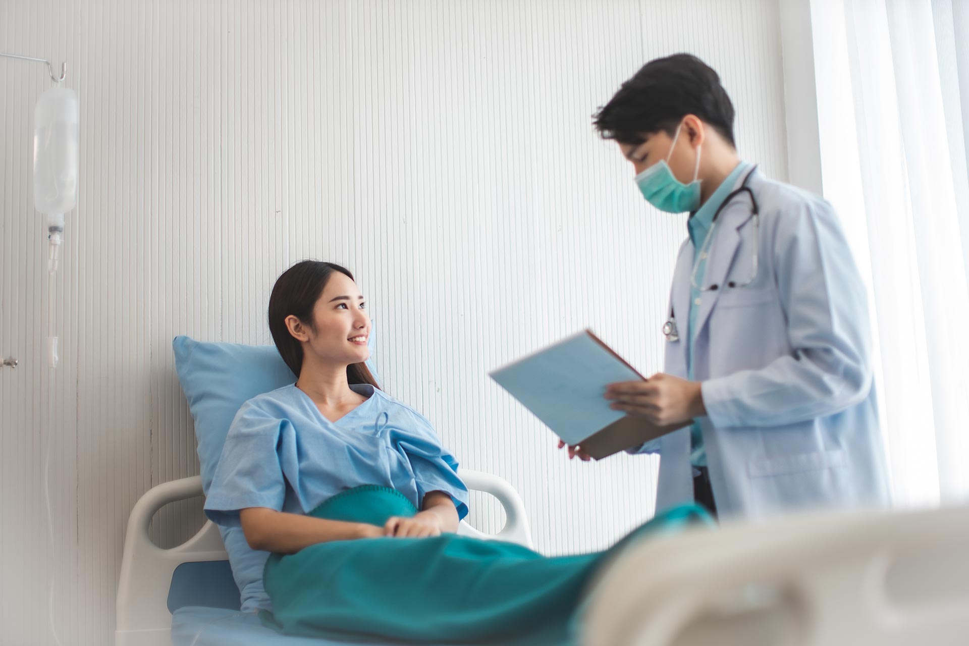 A Vascular Surgeon wearing a mask and white coat stands beside a hospital bed, holding a clipboard, while a patient in a hospital gown lies in bed, smiling and looking at the doctor after a successful vascular surgery. - ARIA Vascular | vascular surgery and interventional Radiology