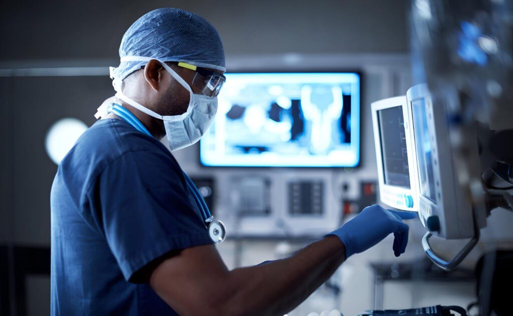 An Interventional Radiology healthcare professional in scrubs, mask, gloves, and hair net operates medical equipment in a dimly lit hospital room, with monitors displaying patient data in the background. - ARIA Vascular | vascular surgery and interventional Radiology